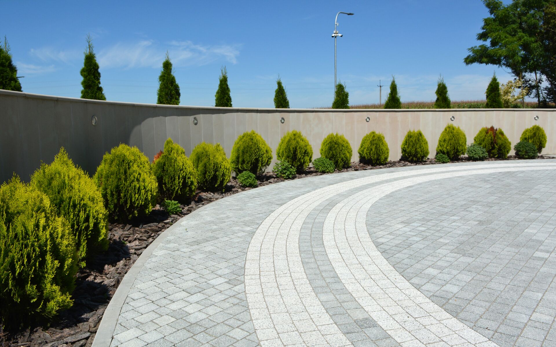 Eleganter, runder Natursteinweg, umgeben von symmetrisch angeordneten Sträuchern und hohen Hecken in einem modernen Garten bei blauem Himmel.