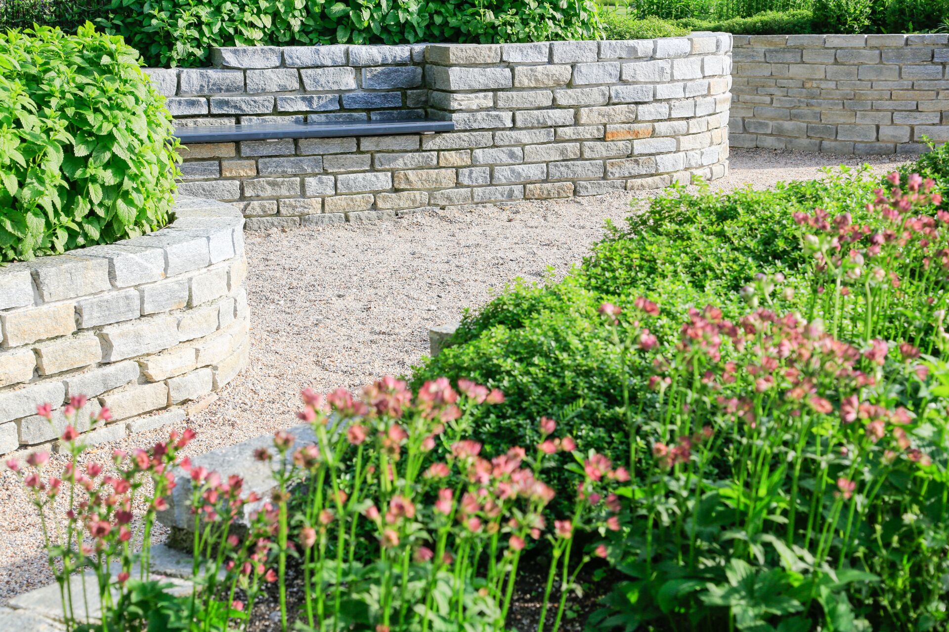Natursteinmauer mit bunten Blumen und grünen Pflanzen in einem sonnigen Garten, umgeben von gepflegten Wegen und einer harmonischen Landschaft.